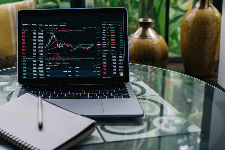 Laptop displaying cryptocurrency stocks and graphs on a glass table with a notepad